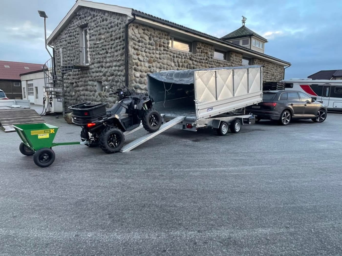 Tilhenger med ATV lastes på ved en steinbygning, med en Audi Q7 bak